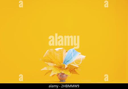 Viele Herbstgelbe Blätter und Gesichtsmaske auf der braunen Vase auf gelbem Hintergrund. Leerraum Bild. Stockfoto