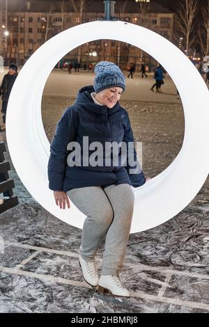 Porträt einer älteren Frau in Schlittschuhen, die am Winterabend auf der Ringbank auf der städtischen Eisbahn ruht. Stockfoto