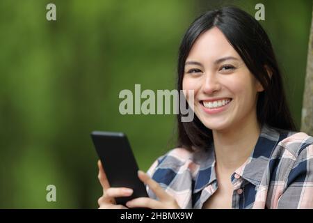 Asiatische Frau lächelt mit Smartphone und schaut in einem Park auf die Kamera Stockfoto