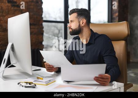 Ein kluger und konzentrierter Kaukasier macht Papierkram. Ein Mann schaute durch Papier und saß am Schreibtisch mit einem Computer im Büro. Man prüft das Berichtskonzept Stockfoto