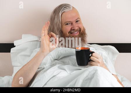 Glücklicher bärtiger Mann, der morgens Kaffee in einem gemütlichen Bett genießt und ein Schild mit OK zeigt. Lächelnder Kerl, der die Kamera anschaut. Nahaufnahme Stockfoto