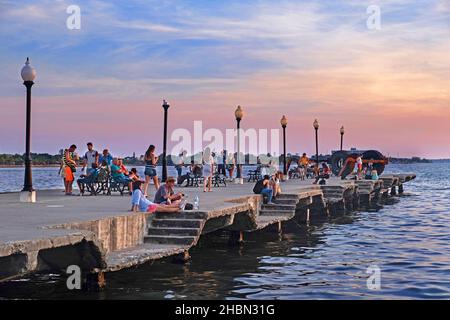 Kubaner und Touristen sitzen auf dem Beton Muelle Real / Royal Pier bei Sonnenuntergang in der Stadt Cienfuegos auf der Insel Kuba, Karibisches Meer Stockfoto