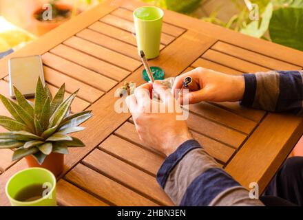 Unerkannte Person an einem Holztisch, die Cannabis-Joint an einem schönen Ort voller Pflanzen raucht. Stockfoto