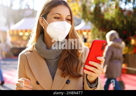 Schöne junge Frau trägt medizinische Gesichtsmaske mit Smartphone in der Weihnachtszeit im Freien Stockfoto