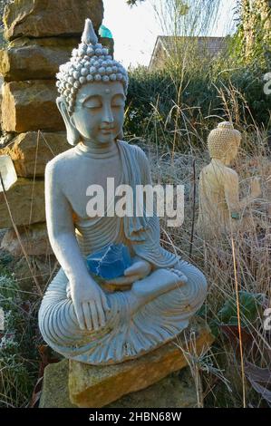 Eine blaue Buddha-Statue mit einem Stück blauem Glas auf seinem Schoß, bedeckt mit Reif. Im Hintergrund ein weißer Buddha Stockfoto