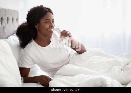Lächelnde, hübsche schwarze Dame, die Wasser im Bett trinkt Stockfoto