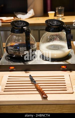 Kaffee- und Milchkannen auf Heizbasis und Brotmesser auf Holzbrett im Frühstücksbereich des Hotels Stockfoto