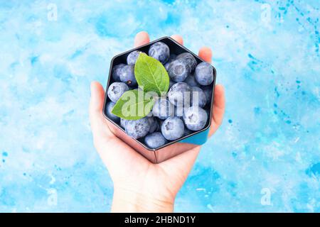 Weibliche Hand hält eine schwarze sechseckige Box mit frischen Blaubeeren und grünen Blättern vor einem hellblauen Aquarellhintergrund. Stockfoto