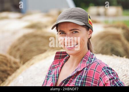 Junge Frau in Hemd und Mütze lächelnd, Heuhaufen rollt hinter ihr Stockfoto