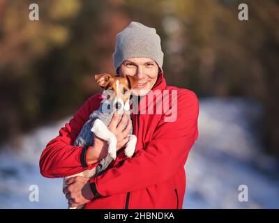 Junger Mann in roter Jacke, der seinen Jack Russell Terrier Hund am Winternachmittag bei den Händen trägt, verschwommener schneebedeckter Pfadhintergrund Stockfoto