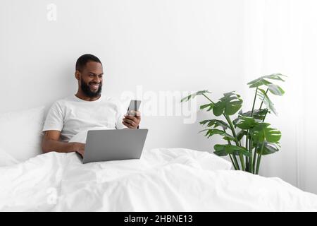 Glücklicher junger schwarzer bärtiger Mann in weißem T-Shirt sitzt auf dem Bett arbeitet auf dem Laptop, liest Nachricht am Telefon Stockfoto