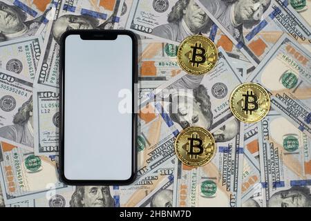 Drei goldene symbolische Bitcoin-Münze auf hundert Dollar Hintergrund mit weißem Bildschirm Smartphone Stockfoto