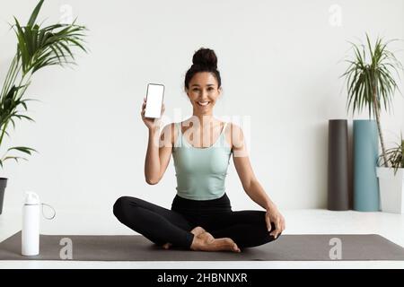 Fitness-App Lächelnde Frau, Die Auf Einer Yogamatte Sitzt Und Leeres Smartphone Zeigt Stockfoto