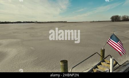 Blick vom Dock des gefrorenen Sees Winneconne mit amerikanischer Flagge im Wind. Am Vortag landete frischer Schnee. Aufgenommen im Dezember 2021. Stockfoto