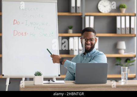 Fröhlicher junger schwarzer Lehrer mit pc-Shows auf der Tafel mit englischen Sprachregeln Stockfoto
