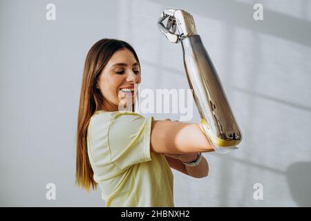 Das Porträt einer starken und unabhängigen Frau mit einem bionischen prothetischen Arm zeigt einen Bizeps auf einem hellen Hintergrund und blickt auf die Kamera und lächelt Stockfoto