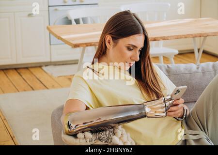 Eine glückliche Frau mit einem bionischen prothetischen Arm sitzt mit einem Telefon auf dem Sofa und kommuniziert online mit Freunden oder surft im Internet oder kauft online in einem gelben T-Shirt vor dem Hintergrund einer hellen Küche ein Stockfoto