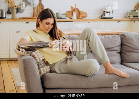 Eine glückliche Frau mit einem bionischen prothetischen Arm sitzt mit einem Telefon auf dem Sofa und kommuniziert online mit Freunden oder surft im Internet oder kauft online in einem gelben T-Shirt vor dem Hintergrund einer hellen Küche ein Stockfoto
