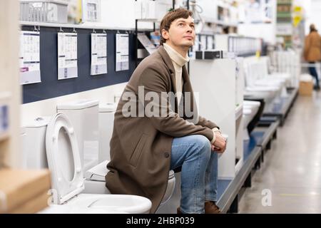 Europäischer Mann in braunem Mantel wählt Toilette und schaut im Baumarkt an die Decke, kauft Toilette für Badezimmer, repariert eine Wohnung Stockfoto