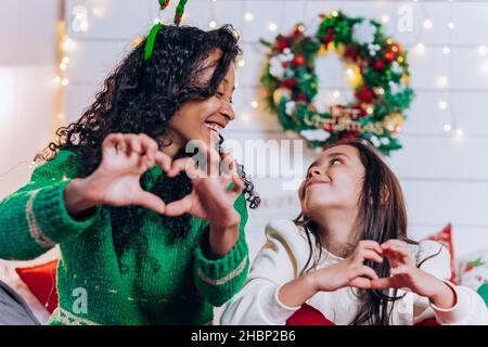 Porträt von niedlichen gemischten Rennen Mutter und Tochter sitzen auf dem Bett zeigen Herzen mit Händen und Blick auf einander vor dem Hintergrund von weihnachtskranz und Girlanden Stockfoto