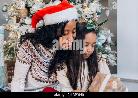 Lockige, langhaarige afroamerikanische Mutter und brünette Tochter sitzen auf dem Teppich und öffnen festliche Geschenke, die in der Nähe des Weihnachtsbaums fröhlich lächeln Stockfoto