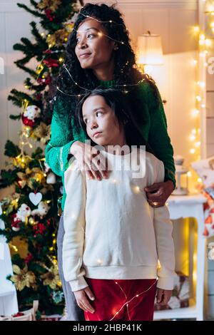 Weihnachtsporträt einer Mutter und Tochter gemischter Rassen am Fenster eines in Girlanden gehüllten Hauses vor dem Hintergrund eines Weihnachtsbaums und von Lichtern Stockfoto