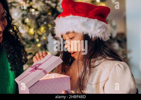 Brünette langhaarige Tochter in Santa Hut öffnet festliche Box mit Geschenken sitzen in der Nähe afroamerikanische Mutter gegen Weihnachtsbaum Stockfoto