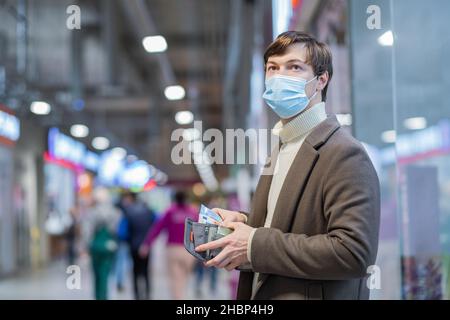 Ein junger Mann in einer medizinischen Maske und einem braunen Mantel legt Euroscheine in eine Brieftasche in einem Einkaufsmarkt, Nahaufnahme Porträt Stockfoto