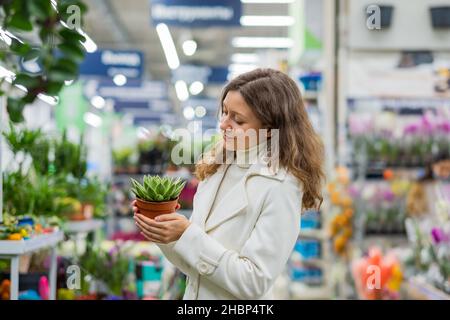 Schöne Frau im weißen Mantel wählt Zimmerpflanze für zu Hause im Blumenladen Stockfoto