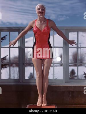 Junge Frau in rot-schwarzem Badeanzug, die auf einem Sprungbrett eines Außenpools steht und schneebedeckte Berge im Hintergrund zeigt Stockfoto