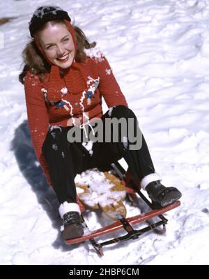Junge Frau im roten Pullover sitzt auf Winterschlitten im Schnee und hat Spaß Stockfoto