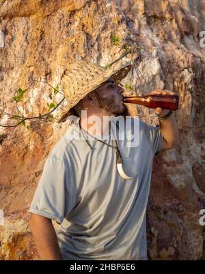 Junger Mann in einem rustikalen Hut, der ein Bier am Strand trinkt. Stockfoto