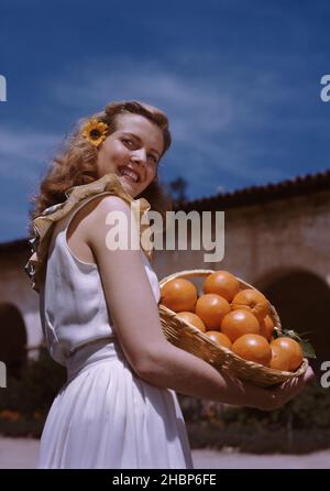Junge blonde Frau mit gelber Gänseblümchen im Haar, in einem weißen Sommerkleid mit einem Korb mit Orangen Stockfoto