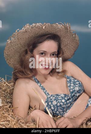 Junge Frau im 2-teiligen blau-weißen Muster-Outfit, die im Heu liegt und einen Strohhut trägt Stockfoto