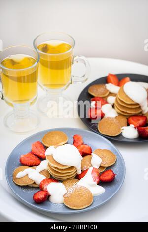 Mini-Pfannkuchen mit saurer Sahnesauce und Erdbeeren auf Tellern, Frühstück für die Familie. Vorderansicht. Stockfoto