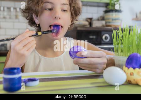 Das Mädchen malt ihre Zunge mit Lebensmittelfarben. Das Mädchen hält einen Kunstpinsel und ein Osterei in der Hand. DIY Ostereier malen. Stockfoto