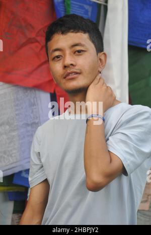 Foto eines asiatischen Mannes, der mit der Hand hinter dem Hals posiert und die Kamera anschaut, der gegen buddhistische Gebetsfahnen steht Stockfoto