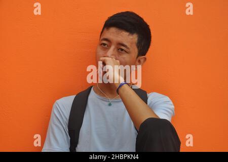 Ein asiatischer junger Mann, der seitwärts schaut, posiert mit Faust auf seinem Gesicht und sitzt vor orangefarbenem Wandhintergrund mit Kopierraum Stockfoto
