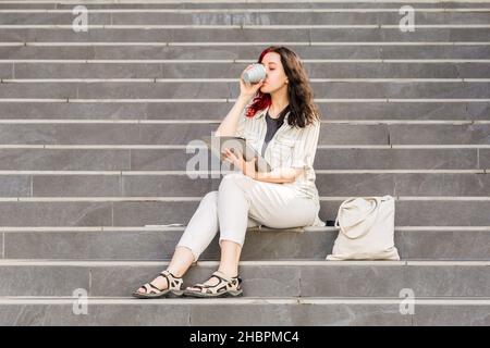 Schöne weiße junge Geschäftsfrau hält Digital Tablet im Freien sitzen auf der Treppe während ihrer Kaffeepause. Kaffee aus wiederverwendbarem Koll trinken Stockfoto