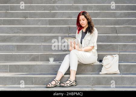 Schöne weiße junge Geschäftsfrau mit Digital Tablet im Freien sitzen auf der Treppe während ihrer Kaffeepause. Wiederverwendbare Kaffeetasse und Leinenshoppi Stockfoto