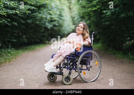 Angenehme junge Dame mit spinaler Muskelatrophie, die einen warmen Sommertag an der frischen Luft verbringt. Kaukasische Frau im Rollstuhl posiert zwischen grünen Park. Stockfoto