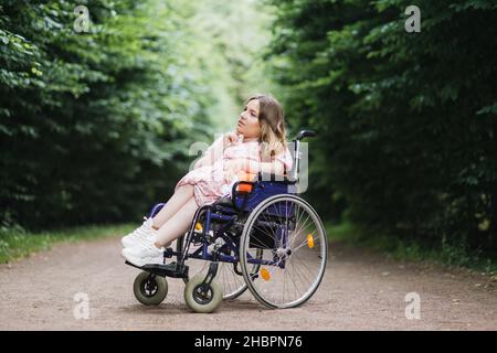 Weibliche Person mit spinaler Muskelatrophie verbringt Zeit allein im Freien mit ihren Gedanken. Junge Frau, die im Rollstuhl sitzt im grünen Sommerpark und schaut zur Seite. Stockfoto