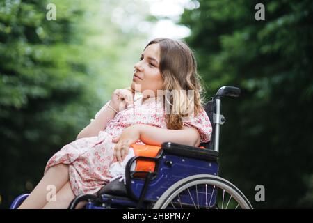 Weibliche Person mit spinaler Muskelatrophie verbringt Zeit allein im Freien mit ihren Gedanken. Junge Frau, die im Rollstuhl sitzt im grünen Sommerpark und schaut zur Seite. Stockfoto