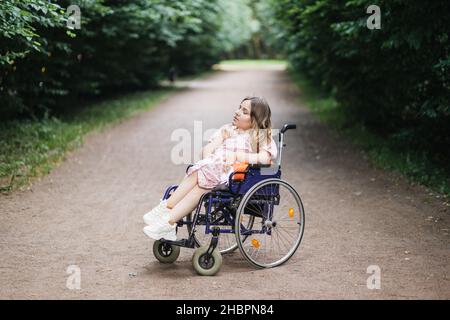 Weibliche Person mit spinaler Muskelatrophie verbringt Zeit allein im Freien mit ihren Gedanken. Junge Frau, die im Rollstuhl sitzt im grünen Sommerpark und schaut zur Seite. Stockfoto