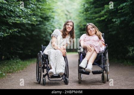 Glückliche junge Frauen mit körperlicher Behinderung genießen Sommerspaziergang im grünen Park. Rollstuhlfahrerinnen, die sich gegenseitig unterstützen und verstehen. Stockfoto