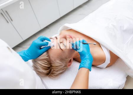 Professionelle Hautpflege im Schönheitssalon. Der Kosmetologe führt eine tiefe mechanische Reinigung mit Spatel des Gesichts des Kunden durch. Blick von den Schultern. Stockfoto