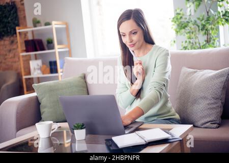 Foto von süßen nachdenklich reifen Dame tragen teal Pullover lächelnd sitzend Couch Schreiben suchen moderne Gadget drinnen Hauszimmer Stockfoto