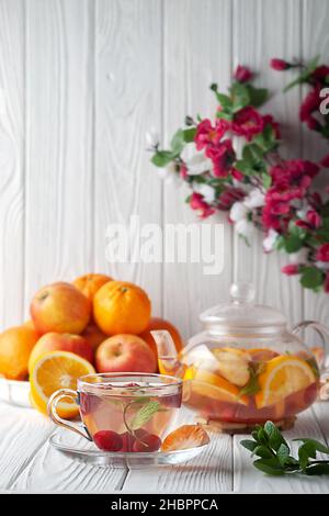 Früchtetee mit Beeren, Orangen und Minze in einer Glas-Teekane auf weißem Holzhintergrund Stockfoto