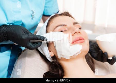 Kosmetologie. Kosmetologe, der mit einer Spatel-Algenmaske auf das lächelnde Gesicht des Weibchens applyiert. Konzept der professionellen Hautpflege. Stockfoto