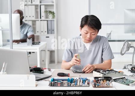 Junge Chinesin in Uniform, die kleine Details der Leiterplatte fixiert, während sie am Schreibtisch gegen ihren afrikanischen männlichen Kollegen sitzt Stockfoto